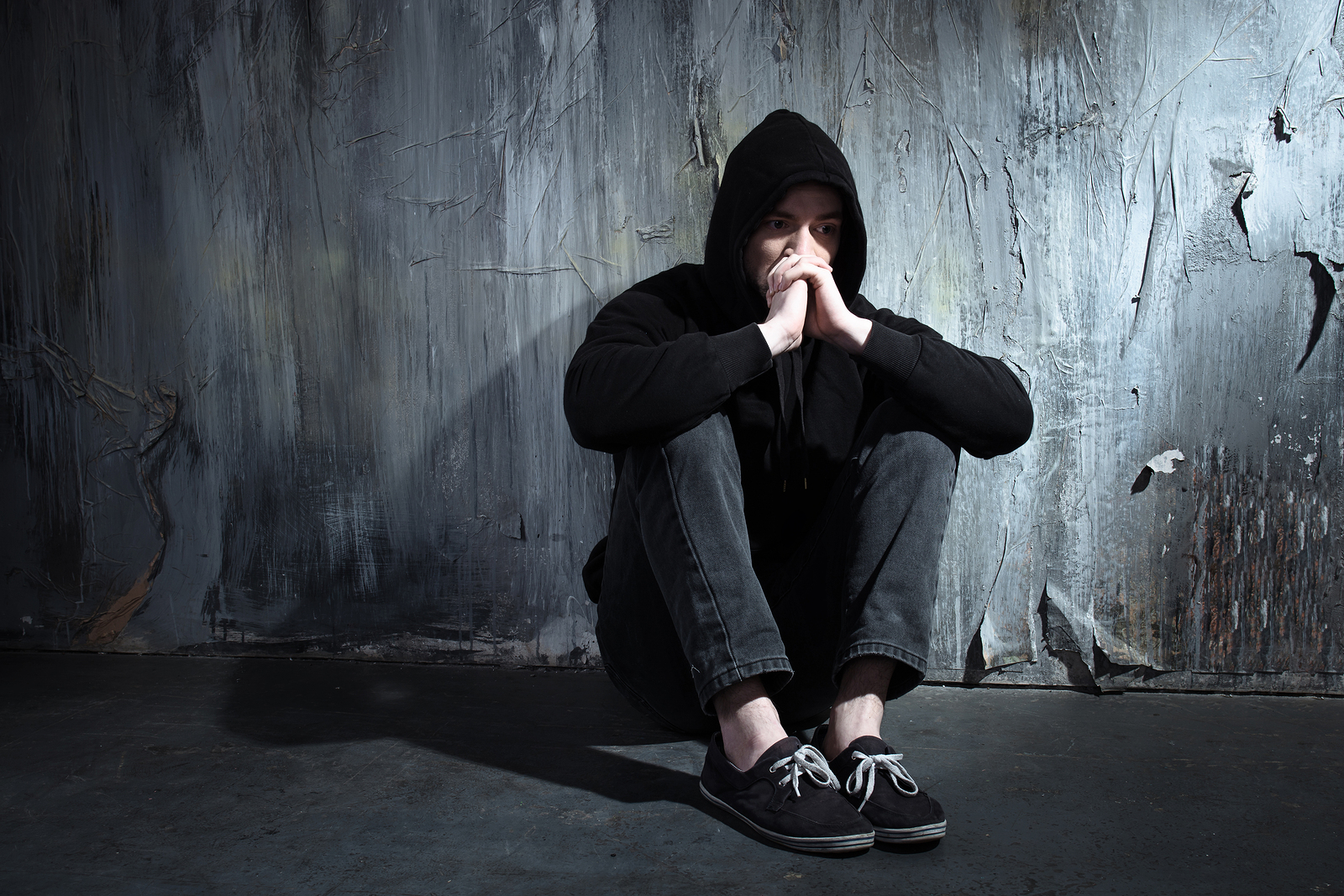 Photo of desperate young drug addict wearing hood and sitting alone in dark 1600 x 1067