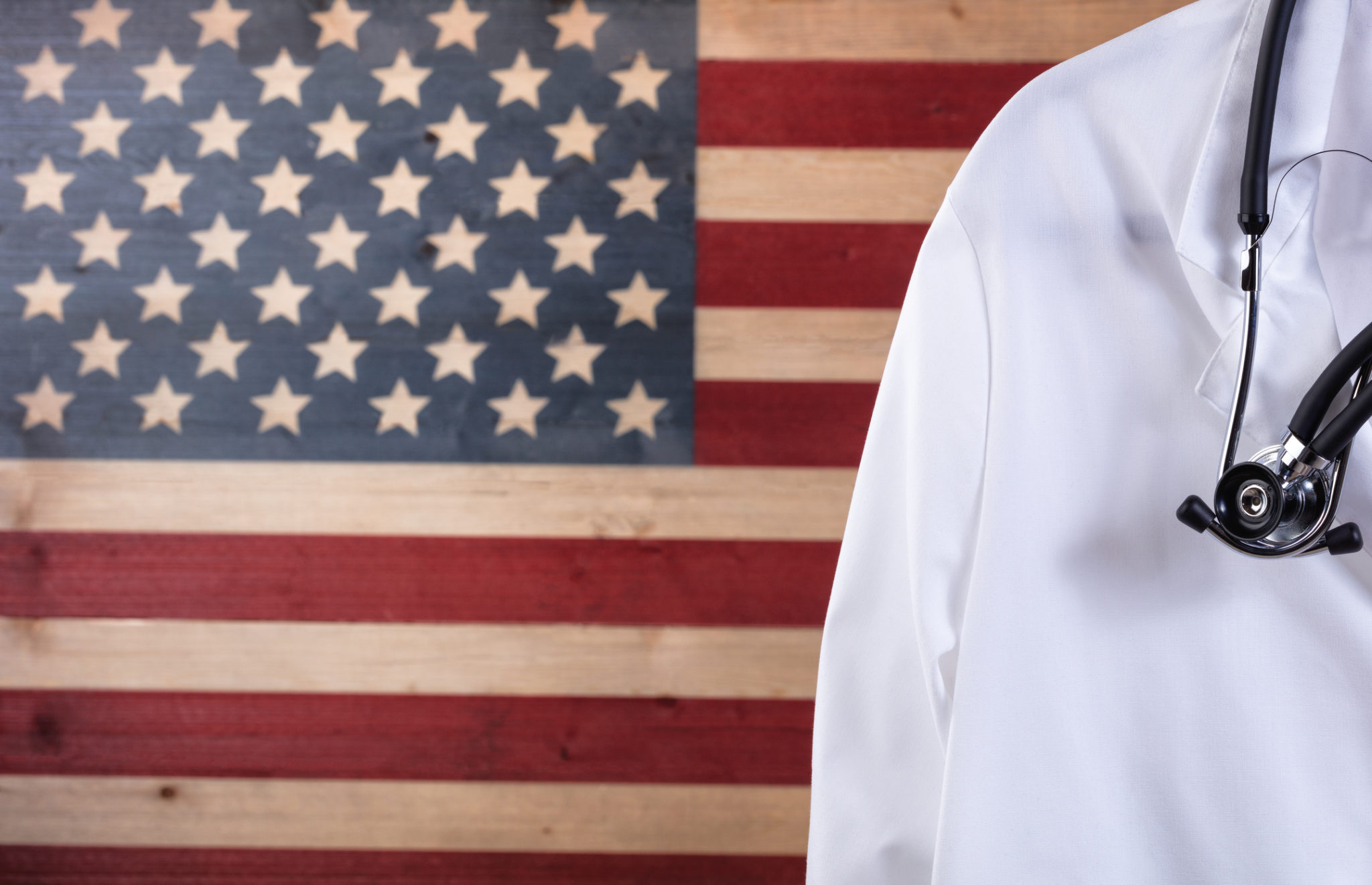 Closeup of medical doctor jacket with stethoscope against faded boards painted in USA flag background. Healthcare concept for America.2048 x 1322